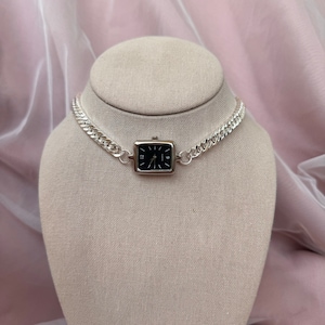 Puede incluir: Un collar choker de cadena plateada con una esfera de reloj negra cuadrada unida al centro.