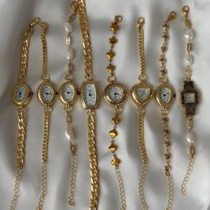 May include: A collection of eight gold-tone wristwatches with white faces and black numerals. The watches feature various band styles, including thick curb chains, delicate heart-shaped links, pearl accents, and small purple circular stones.