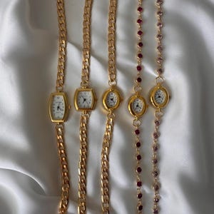 Puede incluir: Cinco relojes de pulsera dorados con esferas blancas y números y agujas negros. Los relojes tienen diferentes formas de esfera, incluyendo rectangulares y ovaladas. Dos de los relojes tienen una cadena de piedras preciosas rojas y rosas.