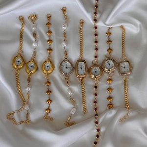 K&ouml;nnte beinhalten: Eine Auswahl goldfarbener Armbanduhren mit verschiedenen Designs. Einige haben ovale oder rechteckige Zifferbl&auml;tter mit wei&szlig;en Ziffernbl&auml;ttern und schwarzen Ziffern. Verzierungen umfassen Perlen, herzf&ouml;rmige Glieder und rote Edelsteine. Die Armb&auml;nder sind auf wei&szlig;em, seidigem Stoff ausgestellt.