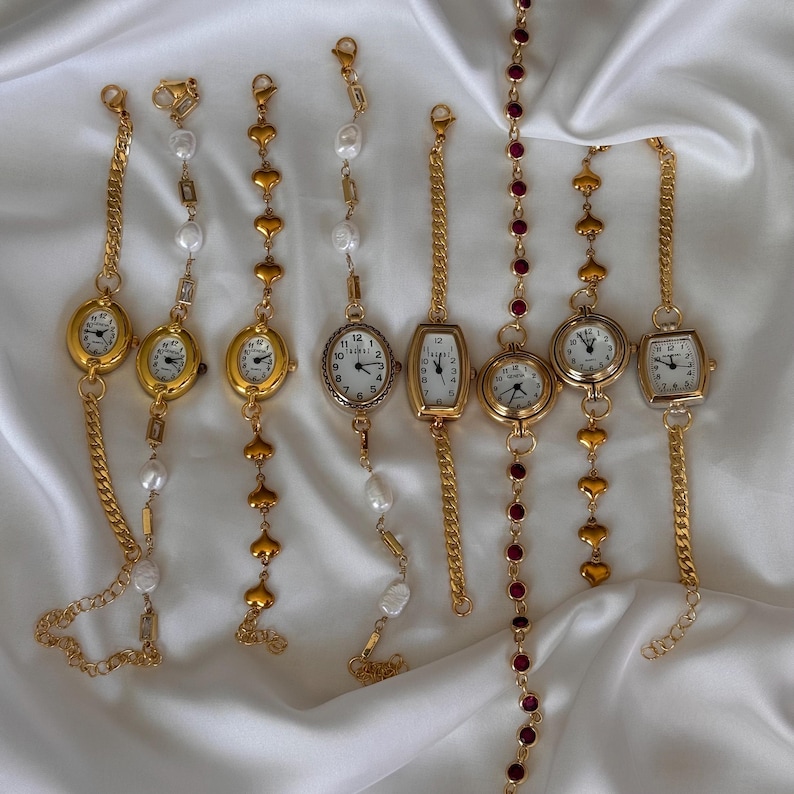 K&ouml;nnte beinhalten: Eine Auswahl goldfarbener Armbanduhren mit verschiedenen Designs. Einige haben ovale oder rechteckige Zifferbl&auml;tter mit wei&szlig;en Ziffernbl&auml;ttern und schwarzen Ziffern. Verzierungen umfassen Perlen, herzf&ouml;rmige Glieder und rote Edelsteine. Die Armb&auml;nder sind auf wei&szlig;em, seidigem Stoff ausgestellt.