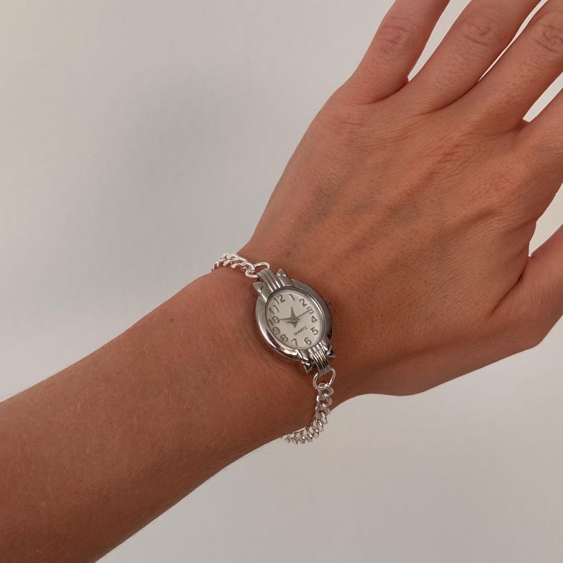 May include: A silver-toned quartz watch with a white face and black numerals. The watch has a chain-link band and is worn on a wrist. The watch face displays the numbers 1 through 12.