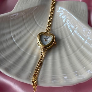 May include: A gold-toned wristwatch with a heart-shaped face and a chain-link band. The watch face is white with black numerals and hands. The watch is displayed on a white, shell-shaped dish, with a pink background.