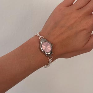 May include: A silver-toned wristwatch with a pink face is worn on a wrist. The watch has a chain-link band. The watch face is oval-shaped and the hands are visible. The watch is a stylish accessory.