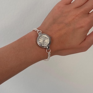 May include: A silver-toned quartz watch with a round face and a chain-link bracelet. The watch face displays the numbers 1 through 12 and has silver hands. The watch is worn on a wrist.