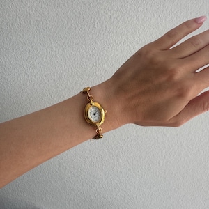 K&ouml;nnte beinhalten: Eine goldfarbene Armbanduhr mit ovalem Zifferblatt und wei&szlig;em Zifferblatt. Die Uhr hat ein zartes Kettengliederarmband. Die Uhr befindet sich an einem Handgelenk vor wei&szlig;em Hintergrund. Das Zifferblatt zeigt schwarze Ziffern und Zeiger.