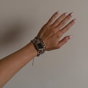 Może przedstawiać: Srebrna bransoletka z łańcuszkiem z kwadratową tarczą zegarka. Tarcza jest czarna z srebrnymi wskazówkami i znacznikami.