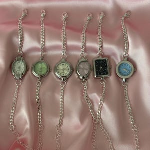 May include: Six silver-toned bracelet watches with different colored watch faces, including white, green, pink, and blue. Each watch has a silver chain and clasp. The watches are displayed on a pink satin surface.