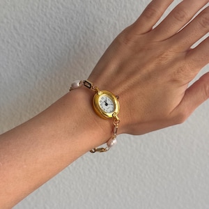 K&ouml;nnte beinhalten: Eine goldfarbene Armbanduhr mit wei&szlig;em Zifferblatt und schwarzen Ziffern wird am Handgelenk getragen. Die Uhr ist mit einem Armband aus Perlen und goldfarbenen Gliedern verbunden. Das Zifferblatt zeigt die Uhrzeit an.