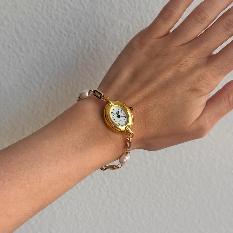 K&ouml;nnte beinhalten: Eine goldfarbene Armbanduhr mit wei&szlig;em Zifferblatt und schwarzen Ziffern wird am Handgelenk getragen. Die Uhr ist mit einem Armband aus Perlen und goldfarbenen Gliedern verbunden. Das Zifferblatt zeigt die Uhrzeit an.