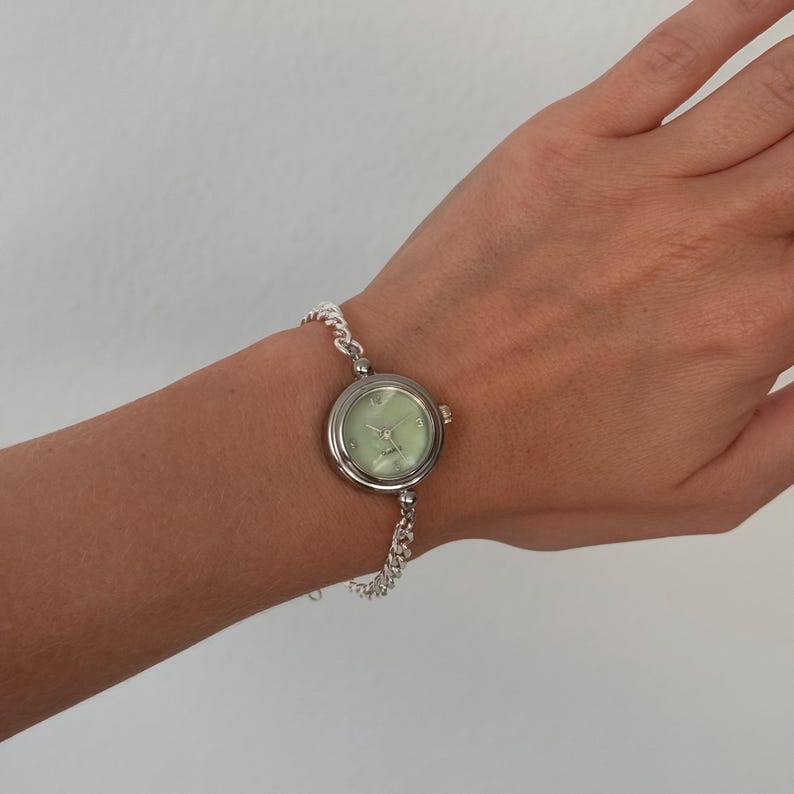 May include: A silver wristwatch with a light green face and silver chain-link band. The watch has a round face with silver hands and hour markers. The watch is worn on a wrist.