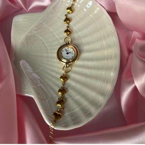 K&ouml;nnte beinhalten: Eine goldfarbene Armbanduhr mit wei&szlig;em Zifferblatt und schwarzen Ziffern. Das Armband der Uhr besteht aus goldenen herzf&ouml;rmigen Gliedern. Die Uhr wird auf einer wei&szlig;en, muschelf&ouml;rmigen Schale vor einem Hintergrund aus rosa Satin pr&auml;sentiert.