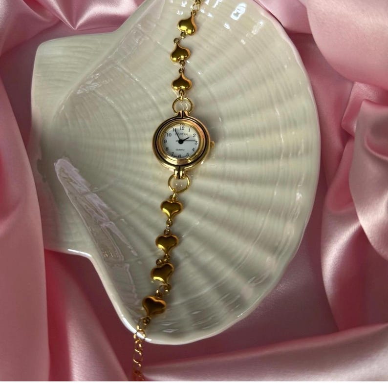 K&ouml;nnte beinhalten: Eine goldfarbene Armbanduhr mit wei&szlig;em Zifferblatt und schwarzen Ziffern. Das Armband der Uhr besteht aus goldenen herzf&ouml;rmigen Gliedern. Die Uhr wird auf einer wei&szlig;en, muschelf&ouml;rmigen Schale vor einem Hintergrund aus rosa Satin pr&auml;sentiert.