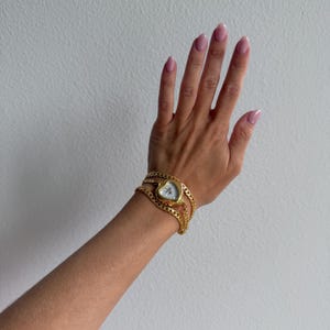 May include: A gold-tone watch featuring a heart-shaped case and a multi-strand chain link bracelet. The watch face is white with gold-tone hands and markers, and the entire piece is constructed from polished gold-colored metal links.