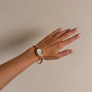 K&ouml;nnte beinhalten: Eine goldfarbene rechteckige Armbanduhr mit wei&szlig;em Zifferblatt und schwarzen Ziffern. Die Uhr ist an einem goldenen Kettenarmband befestigt. Die Uhr befindet sich an einem Handgelenk.
