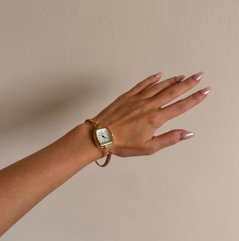 K&ouml;nnte beinhalten: Eine goldfarbene rechteckige Armbanduhr mit wei&szlig;em Zifferblatt und schwarzen Ziffern. Die Uhr ist an einem goldenen Kettenarmband befestigt. Die Uhr befindet sich an einem Handgelenk.