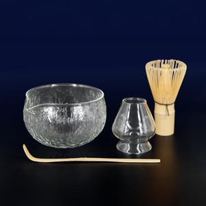 Op de afbeelding: Een matcha-theeset met een getextureerde glazen kom met een tuit, een kleine glazen beker, een bamboe klopper en een bamboe lepel. De set is ontworpen voor het bereiden van traditionele Japanse matcha-thee.