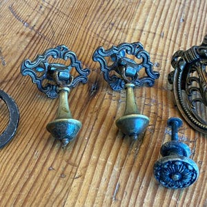 May include: A collection of antique drawer pulls and knobs in various styles. The hardware features ornate designs and a dark bronze finish, with some pieces having brass accents. The pulls include ring pulls and oval-shaped pulls.