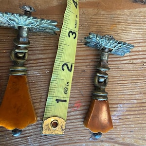 May include: Two antique brass drawer pulls with amber-colored, faceted handles. The pulls have ornate, leaf-like designs and measure approximately 4 inches in length. A yellow measuring tape is placed next to the pulls.