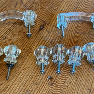 May include: A collection of clear glass cabinet knobs and pulls. The knobs have a faceted design, and the pulls are arched. The hardware is displayed on a wooden surface, showcasing the vintage style.