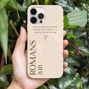 May include: A beige phone case with the text "the pain that you've been feeling, can't compare to the joy that is coming" and "ROMANS 8:18". A small heart symbol is also present. The case is designed for a smartphone.