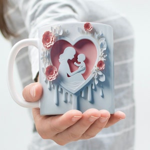 May include: White ceramic mug with a pink heart design featuring a silhouette of a mother holding a baby. The heart is surrounded by pink flowers and white leaves. The mug has a white handle and a blue glaze.