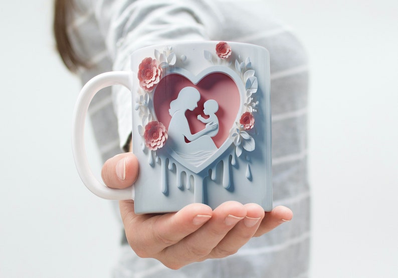 May include: White ceramic mug with a pink heart design featuring a silhouette of a mother holding a baby. The heart is surrounded by pink flowers and white leaves. The mug has a white handle and a blue glaze.