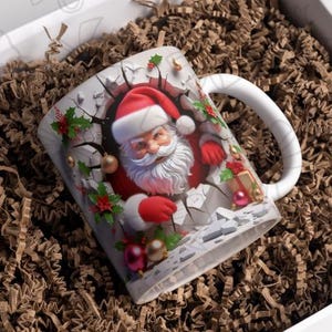 May include: White ceramic mug with a Christmas-themed design featuring Santa Claus breaking through a wall with red and green accents. The mug has a handle and is filled with brown packing peanuts.