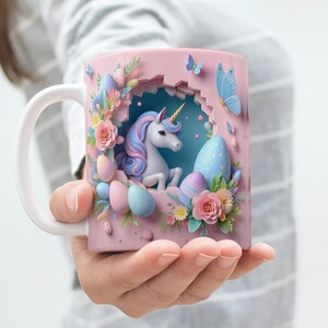 May include: A pink ceramic mug with a 3D design featuring a white unicorn with a blue and pink mane and tail, surrounded by pastel-colored Easter eggs and flowers. The unicorn is peeking out of a hole in the mug.