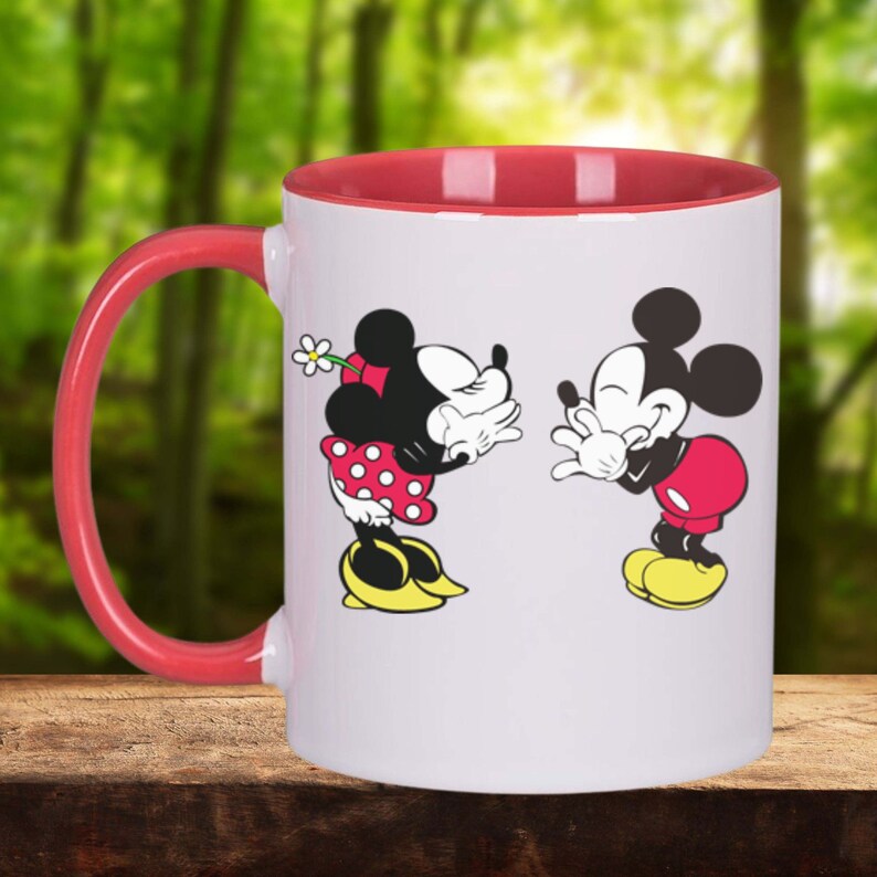 Puede incluir: Una taza de cer&aacute;mica blanca con un borde y un asa rojos. La taza presenta una imagen de dibujos animados de Mickey Mouse y Minnie Mouse bes&aacute;ndose.