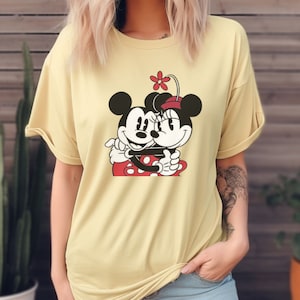 Puede incluir: Una camiseta amarilla con un gr&aacute;fico de Mickey Mouse y Minnie Mouse abraz&aacute;ndose. El gr&aacute;fico es en negro, rojo y blanco.