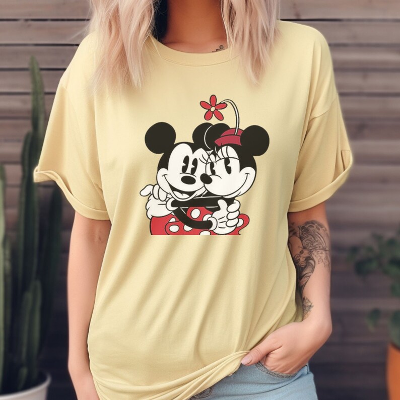Puede incluir: Una camiseta amarilla con un gr&aacute;fico de Mickey Mouse y Minnie Mouse abraz&aacute;ndose. El gr&aacute;fico es en negro, rojo y blanco.