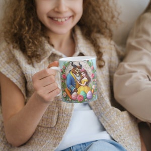 May include: A white ceramic mug with a colorful illustration of Belle and the Beast from Beauty and the Beast. The mug has a blue background with floral accents and pastel-colored Easter eggs.