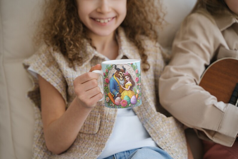May include: A white ceramic mug with a colorful illustration of Belle and the Beast from Beauty and the Beast. The mug has a blue background with floral accents and pastel-colored Easter eggs.