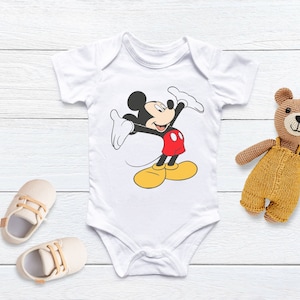 Puede incluir: Un body blanco para beb&eacute; con una imagen de dibujos animados de Mickey Mouse con un atuendo rojo y amarillo. Mickey Mouse est&aacute; saludando con su mano derecha y tiene una gran sonrisa.