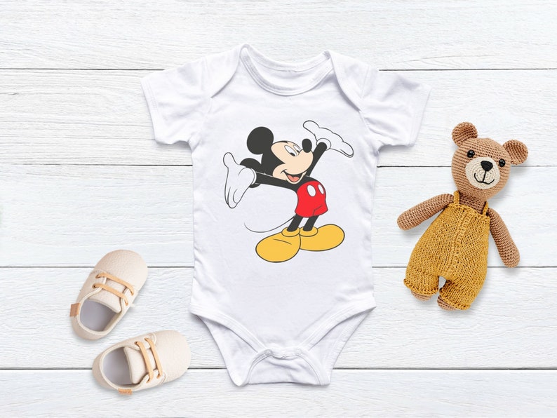 Puede incluir: Un body blanco para beb&eacute; con una imagen de dibujos animados de Mickey Mouse con un atuendo rojo y amarillo. Mickey Mouse est&aacute; saludando con su mano derecha y tiene una gran sonrisa.