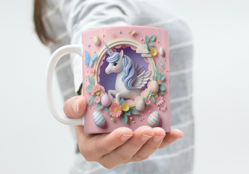 May include: A pink ceramic mug with a white handle features a 3D papercraft design of a unicorn with pastel blue and purple accents. The unicorn is surrounded by flowers and pastel-colored Easter eggs.