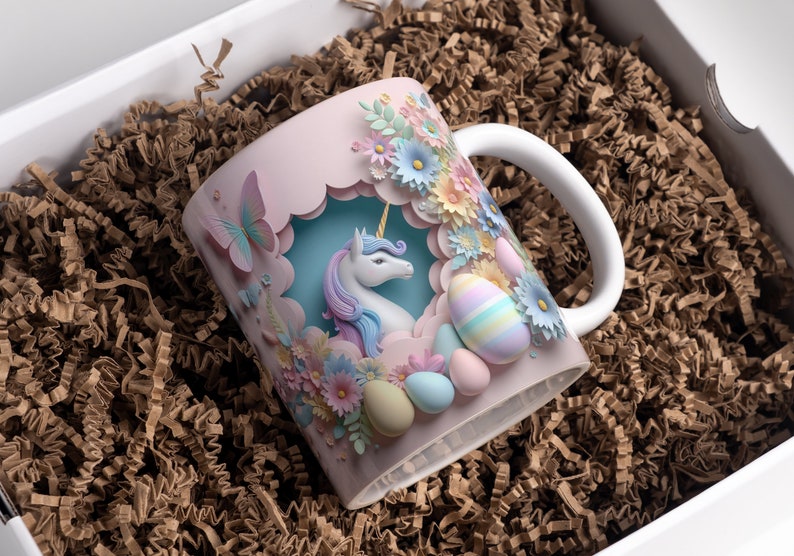 May include: A pink ceramic mug with a 3D paper design featuring a unicorn, flowers, and pastel-colored eggs. The mug has a white handle.