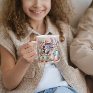 May include: A white ceramic mug with a colorful design featuring a unicorn peeking out of an Easter egg. The mug is surrounded by pastel-colored eggs and flowers.