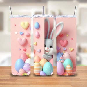 Puede incluir: Tres vasos t&eacute;rmicos rosas y blancos con un tema de Pascua pastel. Cada vaso presenta un conejito de dibujos animados, corazones y huevos coloridos.