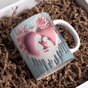 May include: A white ceramic mug with a blue and pink design. The design features a mother and child silhouette inside a heart with pink flowers around the heart. The mug has a white handle.