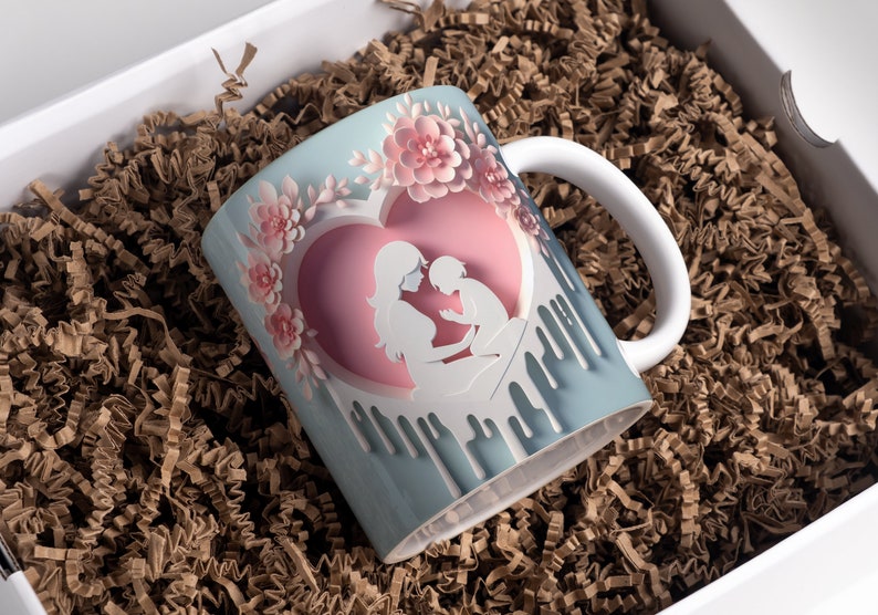 May include: A white ceramic mug with a blue and pink design. The design features a mother and child silhouette inside a heart with pink flowers around the heart. The mug has a white handle.