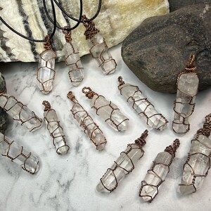 May include: A collection of clear quartz crystal pendants, each wrapped in copper wire. The crystals are elongated and transparent, secured by a decorative wire cage design. Several pendants are attached to black cords, while others are loose.