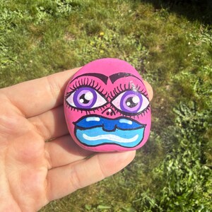 May include: A pink painted rock with a face design. The rock features large purple eyes with long eyelashes, black eyebrows, and blue lips. The rock is held in a hand against a background of green grass.