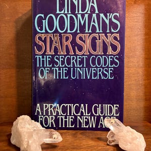 Puede incluir: Un libro de tapa dura titulado Linda Goodman's Star Signs: The Secret Codes of the Universe, A Practical Guide for the New Age, con una cubierta azul oscuro y texto blanco, flanqueado por dos figuras de animales de cuarzo bruto.