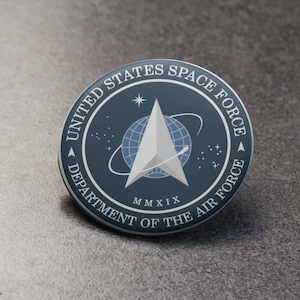 May include: A blue and white round pin with the United States Space Force logo. The logo features a white delta symbol with a globe inside, surrounded by a ring with stars. The text "United States Space Force" is written around the top of the logo, and the text "Department of the Air Force" is written around the bottom of the logo. The year "MMXIX" is written below the delta symbol.