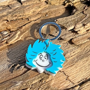 May include: A blue and white keyring featuring a cartoon character with a large smile and blue hair. The keyring has a silver metal ring.