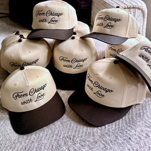 May include: A collection of baseball caps featuring a cream-colored crown and a dark brown brim. Each cap is embroidered with the black cursive text "From Chicago with Love" on the front panel. The hats have a structured design with a button on top.