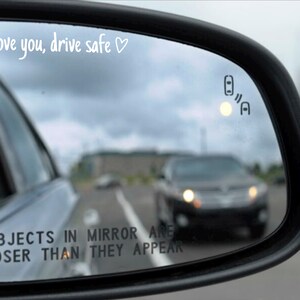 May include: A rearview mirror with the text "I love you, drive safe" written on it. The mirror is showing a car driving in the distance. The text "OBJECTS IN MIRROR ARE CLOSER THAN THEY APPEAR" is also visible on the mirror.