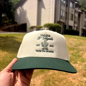 May include: A cream-colored baseball cap with a dark green brim. The front features dark green embroidery of a cactus graphic, the text "Simina & Tequila", "EST 1976", and "Valle De Bravo" centered on the crown of the structured hat.